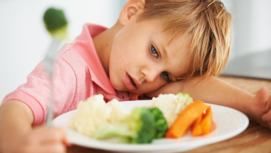 Fair haired boy with his head on one arm with plate of broccoli, carrots and potatoes in front of him