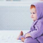 beautiful smiling, nine month old, red-haired infant baby girl in dinosaur costume, is sitting on the bed