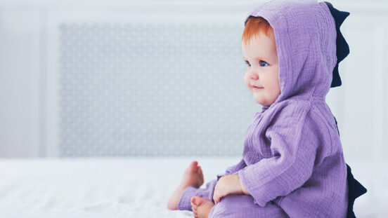 beautiful smiling, nine month old, red-haired infant baby girl in dinosaur costume, is sitting on the bed