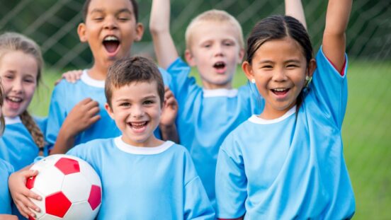 A small group of elementary aged children cheer after a soccer match as they celebrate their win. They are each wearing a blue jersey and are smiling.