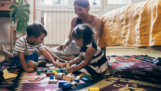 Brother and his little sister sitting on the carpet with their mother, playing with wooden building blocks in their bedroom.