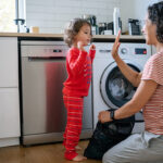 Eurasian three year old girl who is wearing pajamas gives her mom a high-five after helping her do the laundry at home.