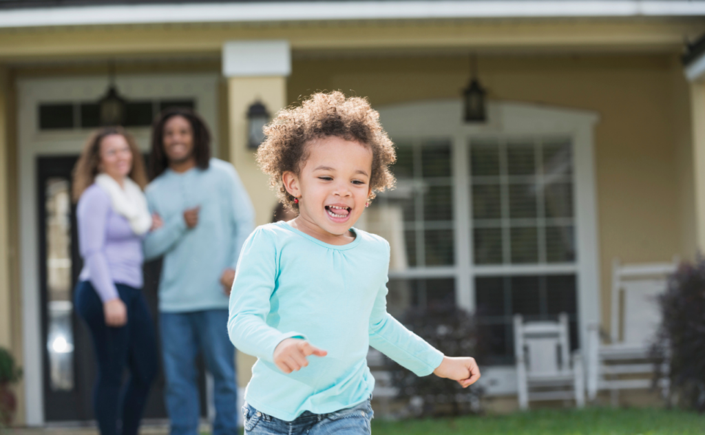 Young girl with dark curly hair happily running in front of her parents standing in front of their house