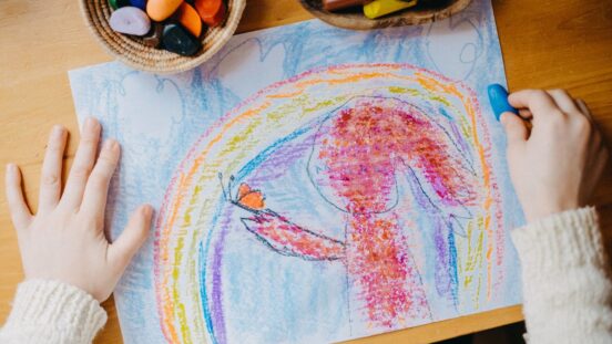 Child's crayon drawing of a rainbow in a blue sky and a pony tailed child with an orange butterful on their hand