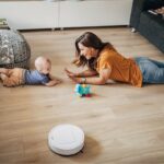 Happy mother playing with her baby son while robot vacuum cleaning floor at home