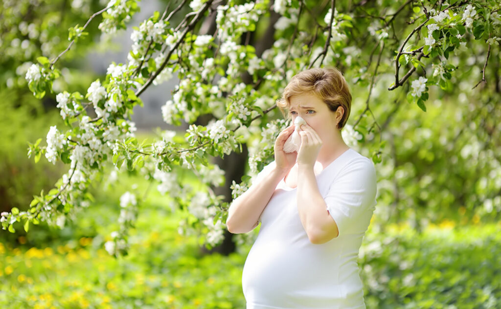Pregnant young woman sneezing and wipes nose with napkin during walking in spring park. Flu season, cold rhinitis
