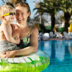 By a blue pool with palm trees and loungers a happy mother is holding a laughing sunglasses wearing son in a big inflatable swim ring