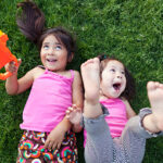 Two toddler girls lie on grass laughing as they play with bubble machine.