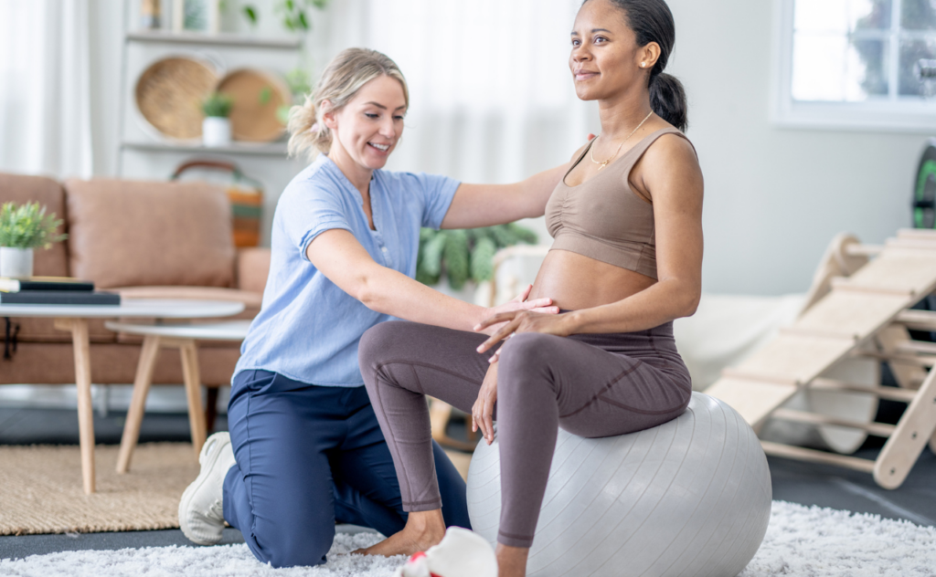 Pregnant woman wearing sports gear, sitting on a yoga ball while a woman in casual scrubs kneels next to her with her hand on her belly