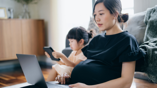 Pregnant Asian woman looking at her laptop while a toddler girl looks at a tablet in the background