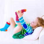 Fair-haired toddler girl wearing brightly coloured clothes lying on her back on a pale sofe with her feet in the air