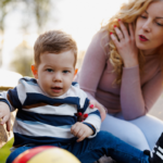Mum sitting on park bench holding a toddler's arm as he looks away not listening to her
