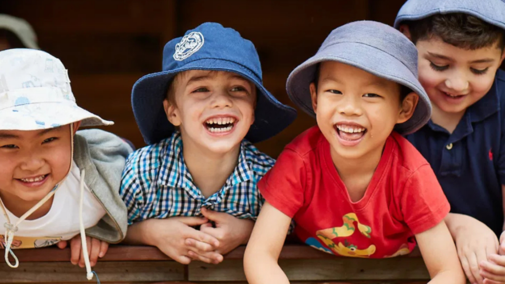 Four smiling children in bucket hats lean forward laughing at the Only About Children Childcare Centre