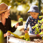 A smiling childcare provider and a young child in a bucket hat are gardening in the sun at Only About Children childcare centre
