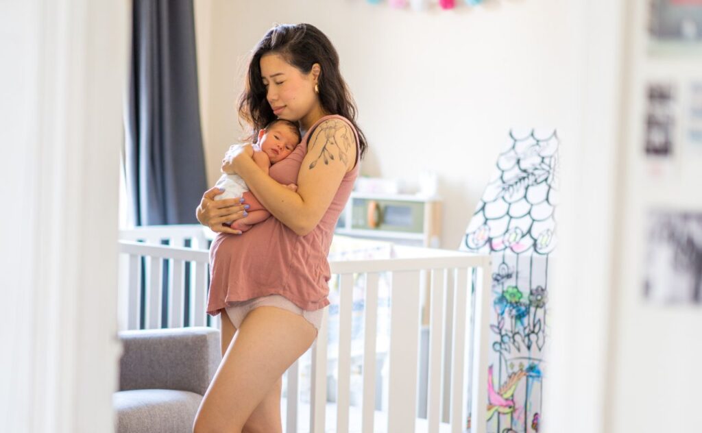 Nursery fan, summer heat: A new Mother stands in her baby's nursery in underwear and a tank top as she recovers at home. She is holding her baby tightly to her chest as she embraces he postpartum phase.