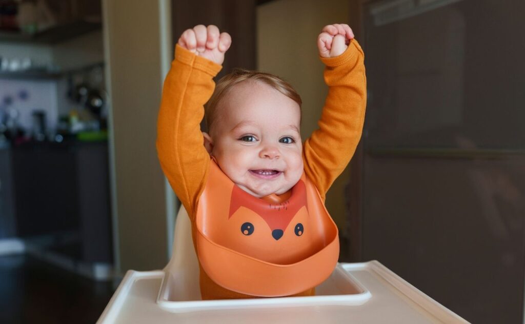 Winning baby name lists: baby boy wearing bib with arms raised sitting on high chair