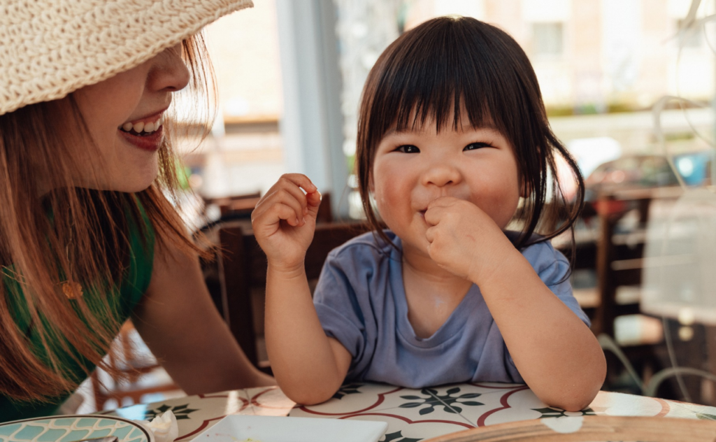 toddler girl smiling and eating while mother smiles at her