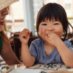 toddler girl smiling and eating while mother smiles at her